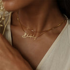 Gouden naamketting met een naam in schrijfletters, gedragen met een tweede dunne ketting (voorbeeldnaam)