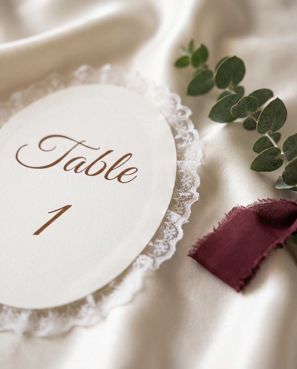 Close-up van een ovale kaart met kantrand op ivoorkleurig satijn naast een bordeaux lint en eucalyptus