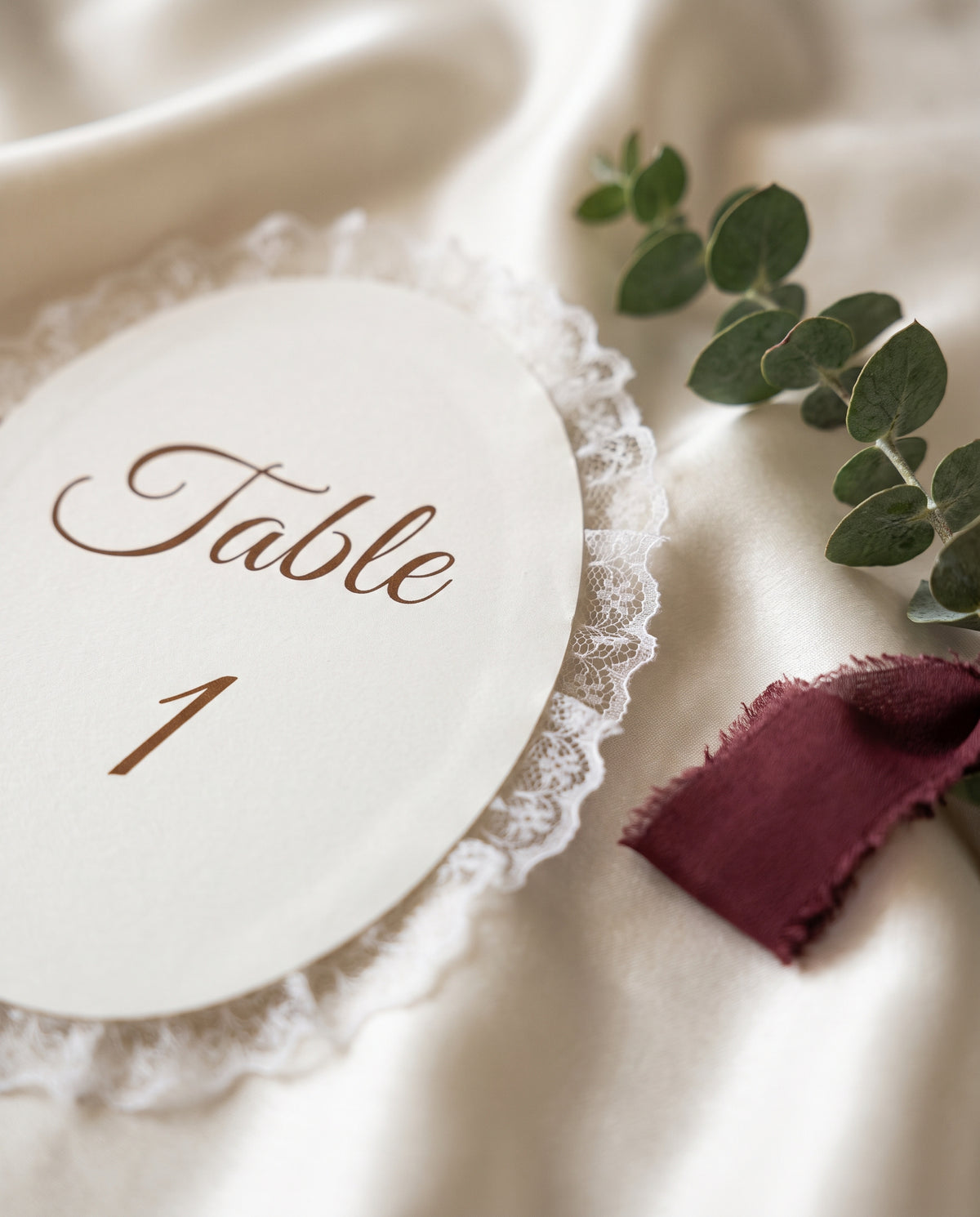 Close-up van een ovale kaart met kantrand op ivoorkleurig satijn naast een bordeaux lint en eucalyptus